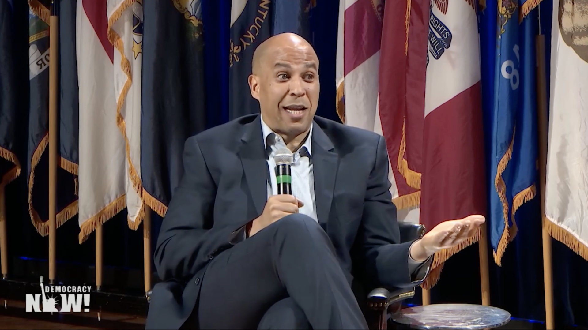 Senator Cory Booker speaks at an event with flags in the background.