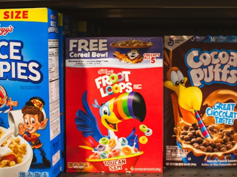 Cereal boxes on a shelf: Rice Krispies, Froot Loops ("Free Cereal Bowl"), and Cocoa Puffs ("Great Chocolate Taste!").