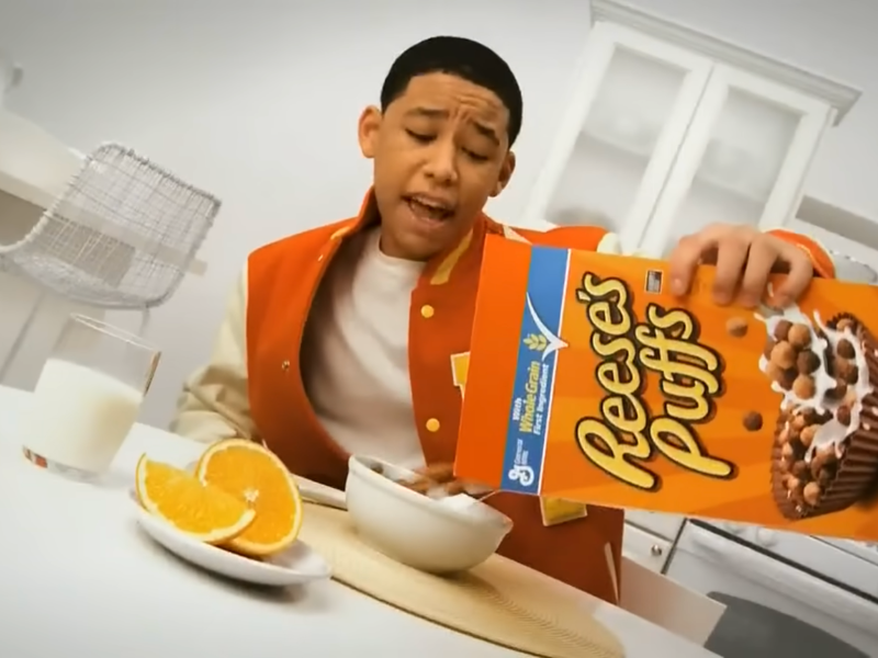 Boy pouring Reese's Puffs cereal into a bowl. Reese's Puffs box visible.