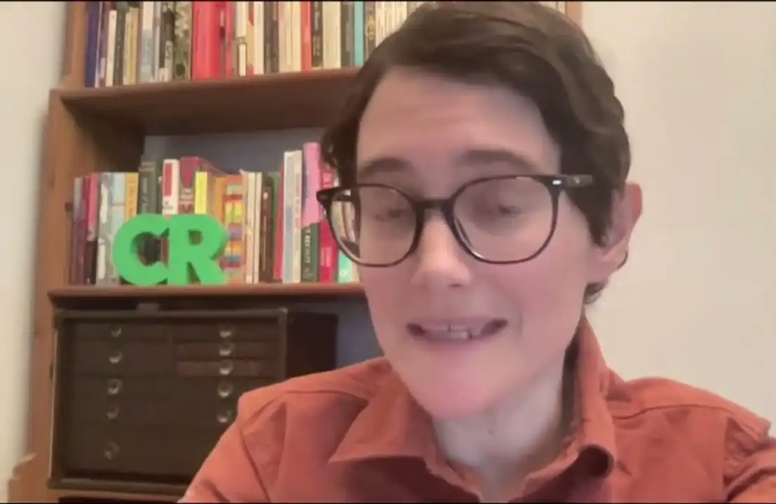Person with glasses in front of a bookshelf and drawers.
