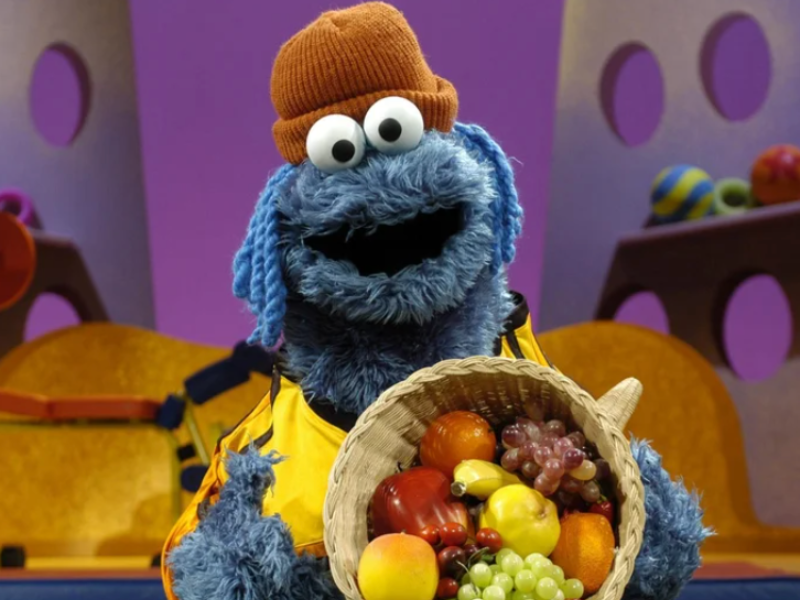 Cookie Monster in orange hat holds basket of diverse fruits for minority marketing.