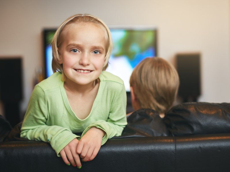 Girl smiling, watching TV with sibling on couch. Home entertainment.