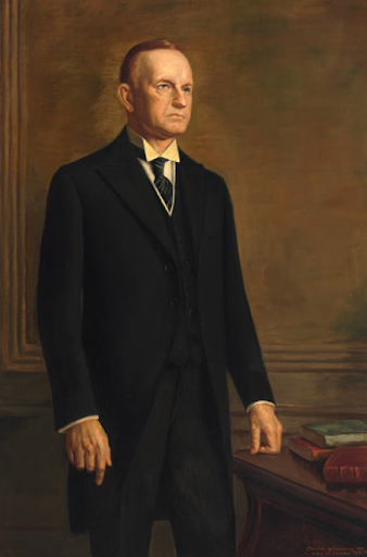 Formal portrait of Calvin Coolidge in a dark suit, standing by a desk.