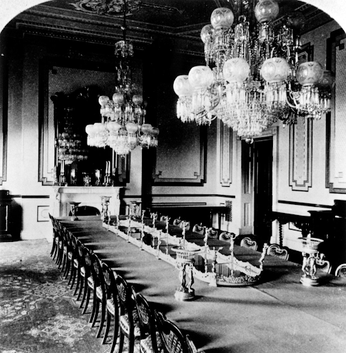 Grand dining room with long table, ornate chandeliers, and fireplace.