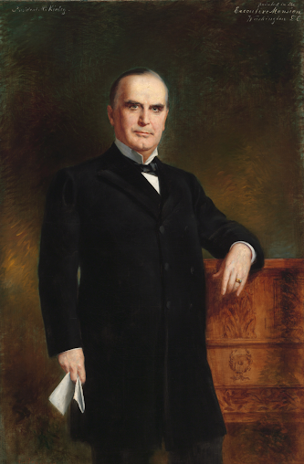 Formal portrait of William McKinley in a black suit, standing next to a wooden cabinet.