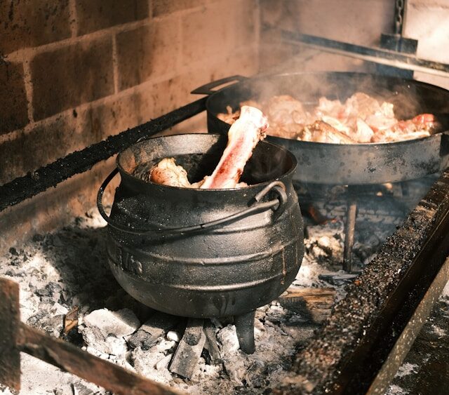 Meat cooking in a traditional cast iron pot over coals in a brick oven.