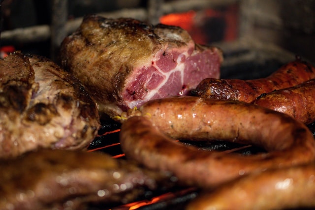 Assortment of grilled meats, including sausages and a large cut of pork, cooking over coals