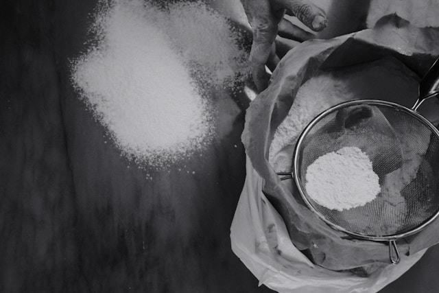 Sifting flour for baking, with a pile of white powder on a dark surface.
