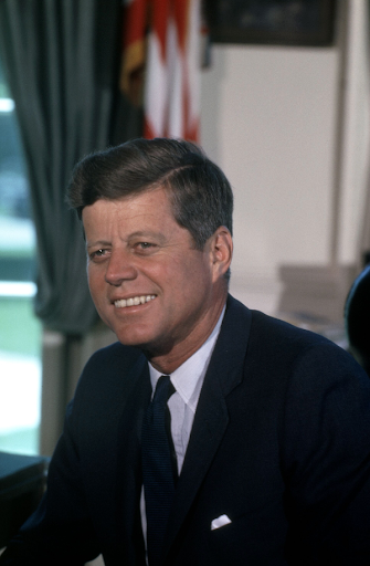 John F. Kennedy in a dark suit and tie, smiling in an office setting.
