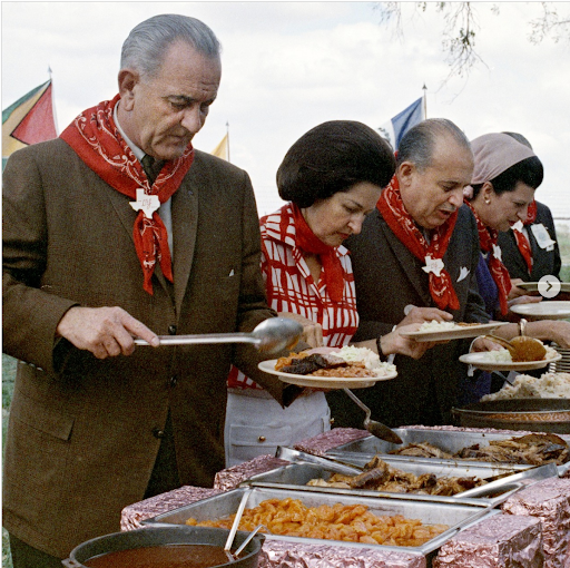Lyndon B. Johnson serves food at a Texas BBQ, wearing a red bandana.