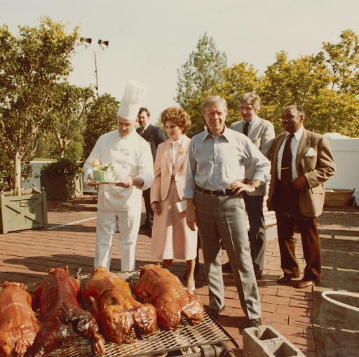 Jimmy Carter and others observe a chef presenting roasted pigs at an outdoor event.