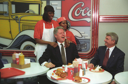 Bill Clinton laughs with two servers at a diner with Coca-Cola memorabilia.