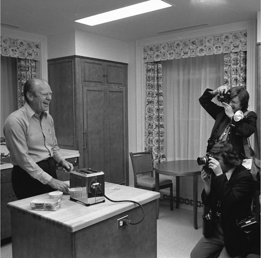Gerald Ford laughs while making toast in a kitchen, photographed by two people.