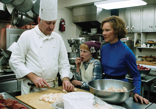 Chef prepares food with Rosalynn Carter and a young girl in a kitchen.