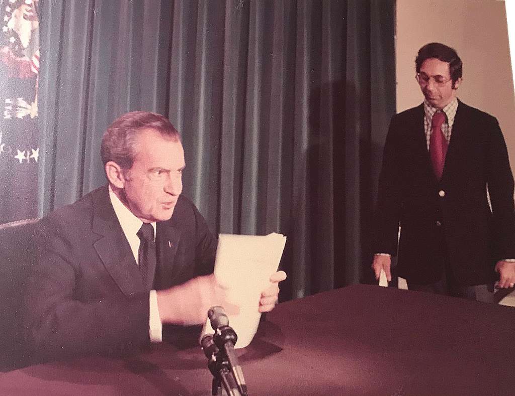 Richard Nixon reading a document at a table with an aide standing behind him.